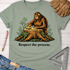 Women's funny woodworking graphic tee shirt for female woodworkers, makers, and DIY builders — Sawdust & Ponytails. Sage green cotton tee with a Bigfoot sanding wood on a tree stump, "Respect the process." text.