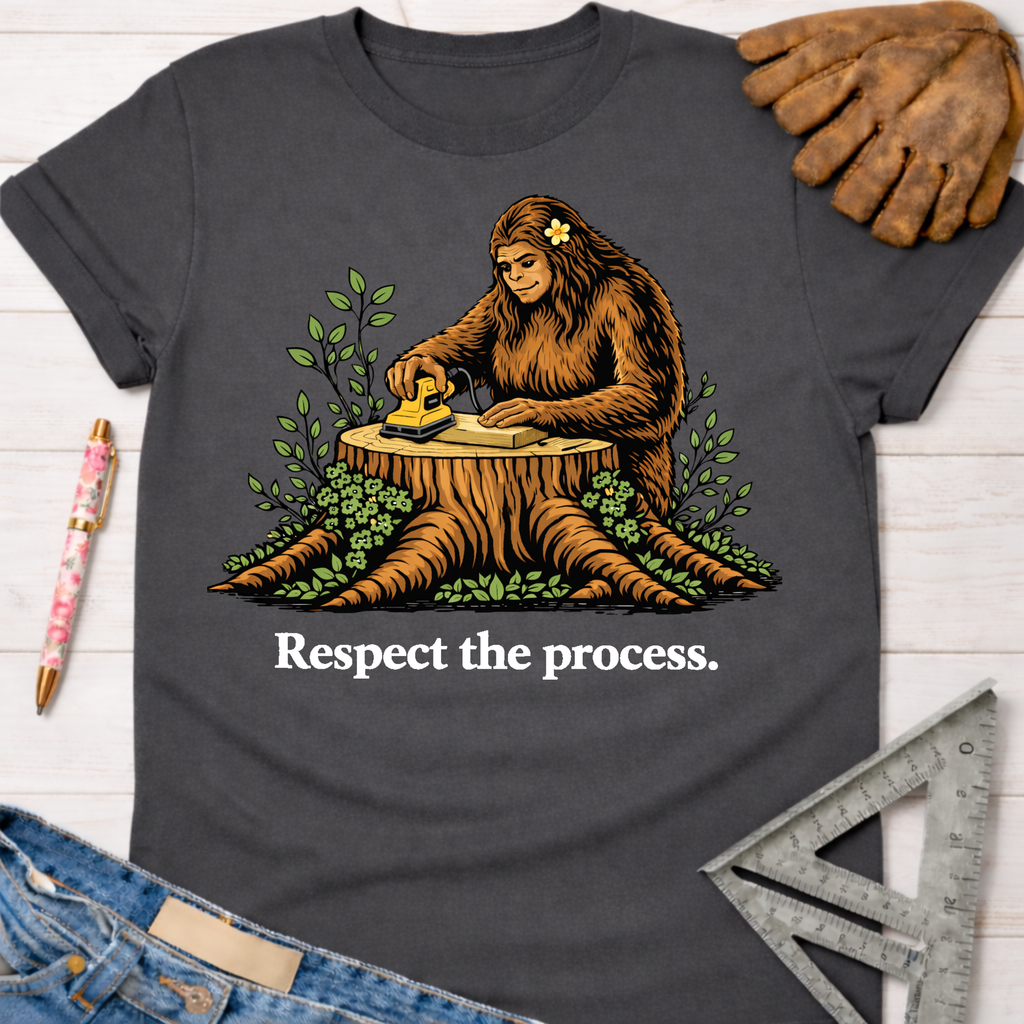 Women's funny woodworking graphic tee shirt for female woodworkers, makers, and DIY builders — Sawdust & Ponytails. Dark gray t-shirt with a Bigfoot sanding wood on a tree stump, text "Respect the process.