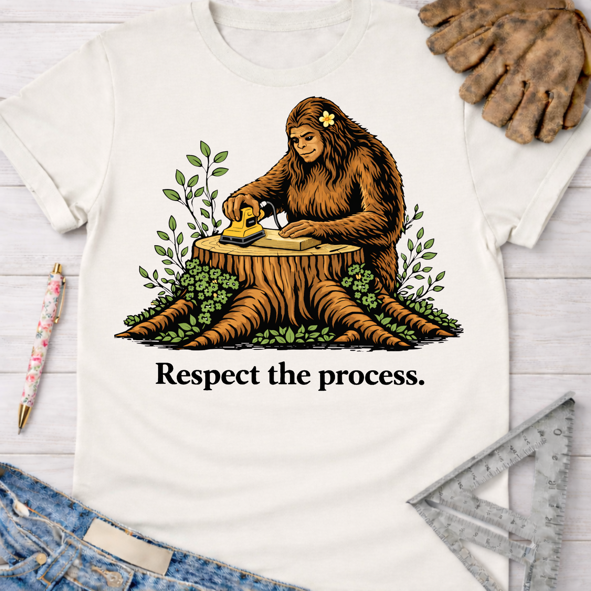 Women's funny woodworking graphic tee shirt for female woodworkers, makers, and DIY builders — Sawdust & Ponytails. White cotton tee with Bigfoot sanding wood on a tree stump, "Respect the process." text.