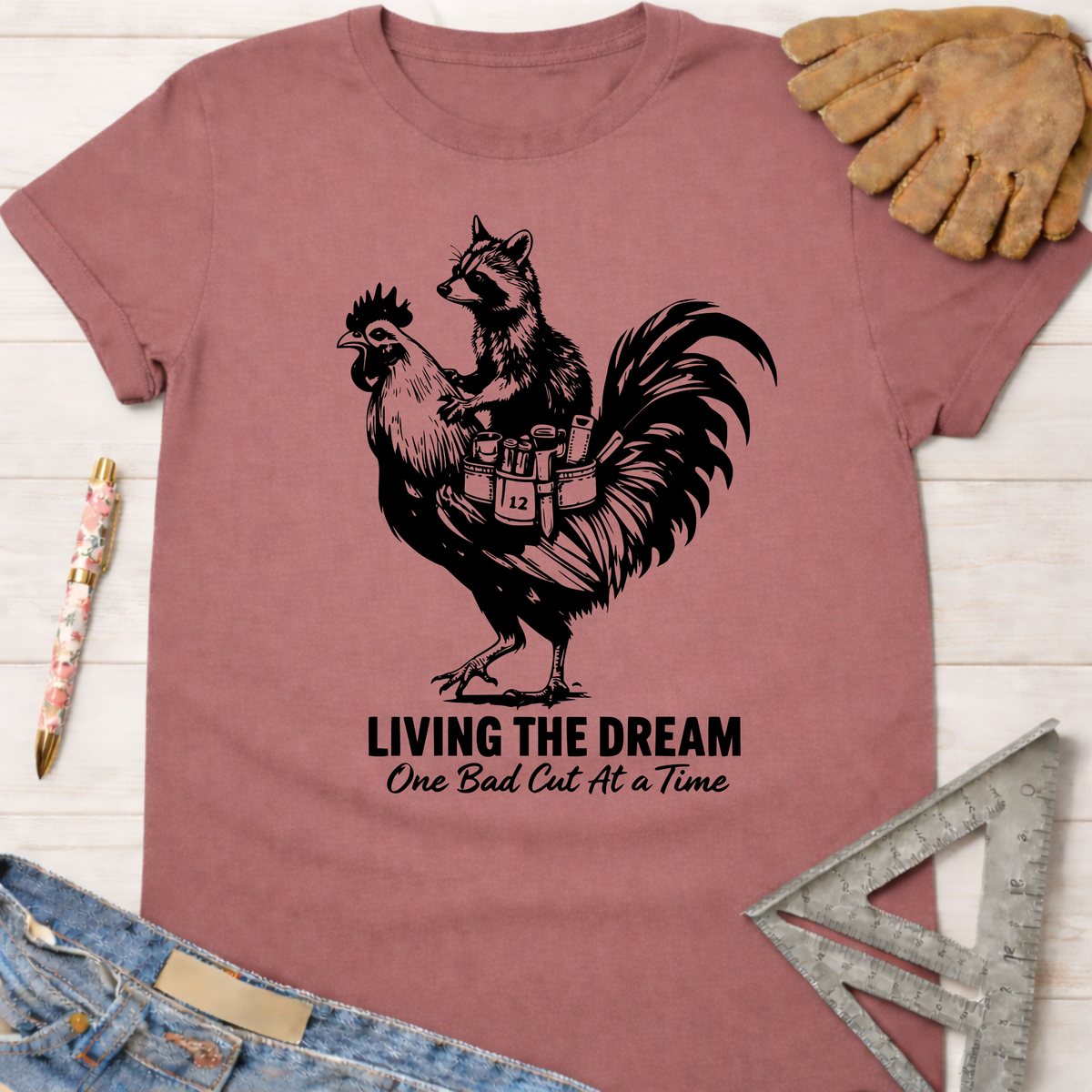 Women's funny woodworking graphic tee shirt for female woodworkers, makers, and DIY builders — Sawdust & Ponytails. Mauve t-shirt with a raccoon riding a rooster, text "LIVING THE DREAM One Bad Cut At a Time".
