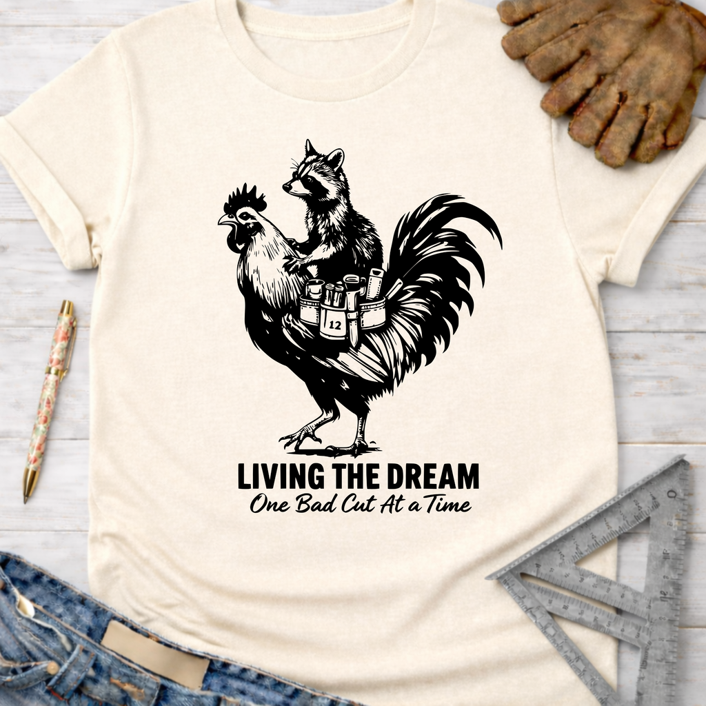 Women's funny woodworking graphic tee shirt for female woodworkers, makers, and DIY builders — Sawdust & Ponytails. Cream unisex tee with a raccoon riding a rooster, "Living The Dream One Bad Cut At A Time" text.