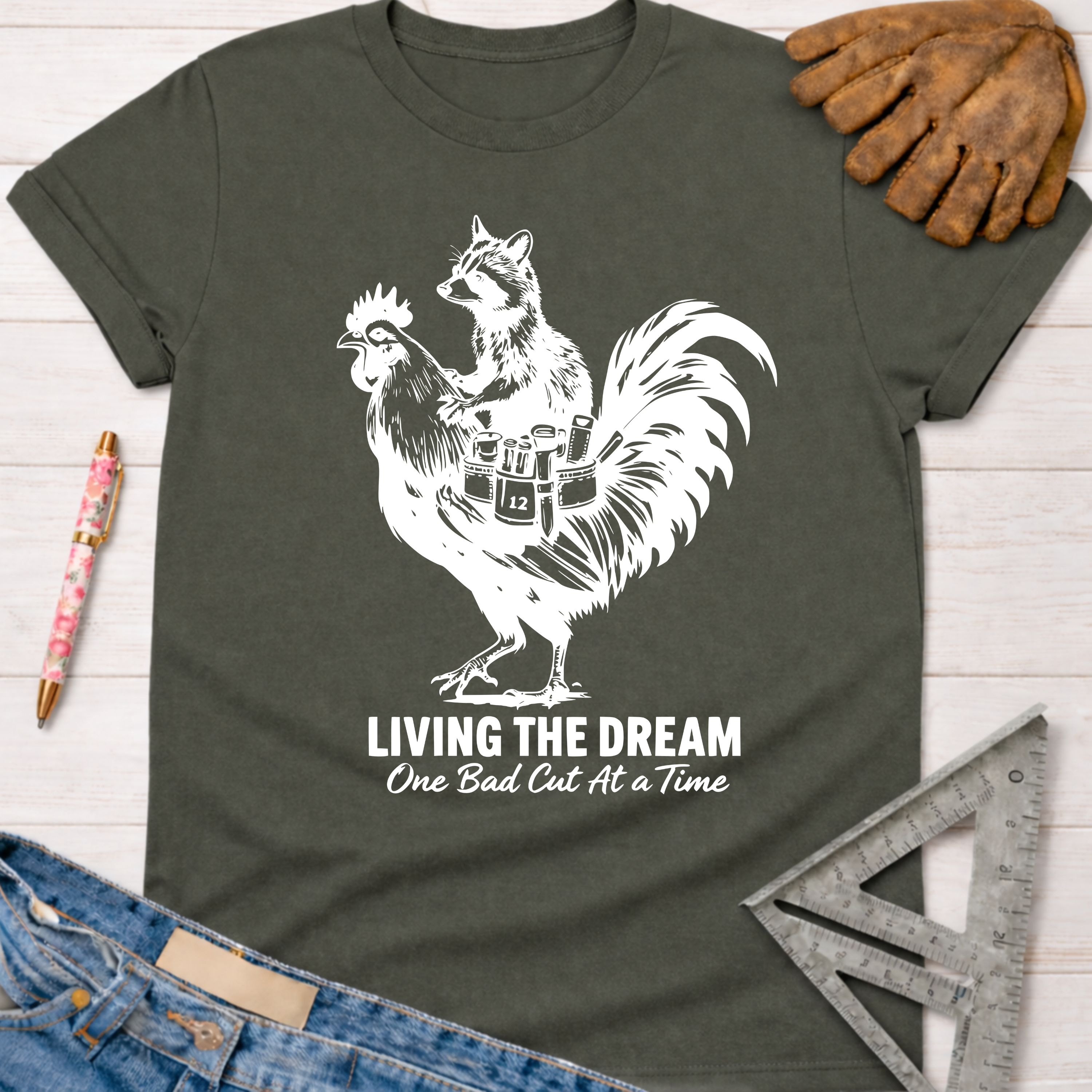 Women's funny woodworking graphic tee shirt for female woodworkers, makers, and DIY builders — Sawdust & Ponytails. Dark gray tee with white graphic of a raccoon riding a rooster, "LIVING THE DREAM One Bad Cut At a Time" text.