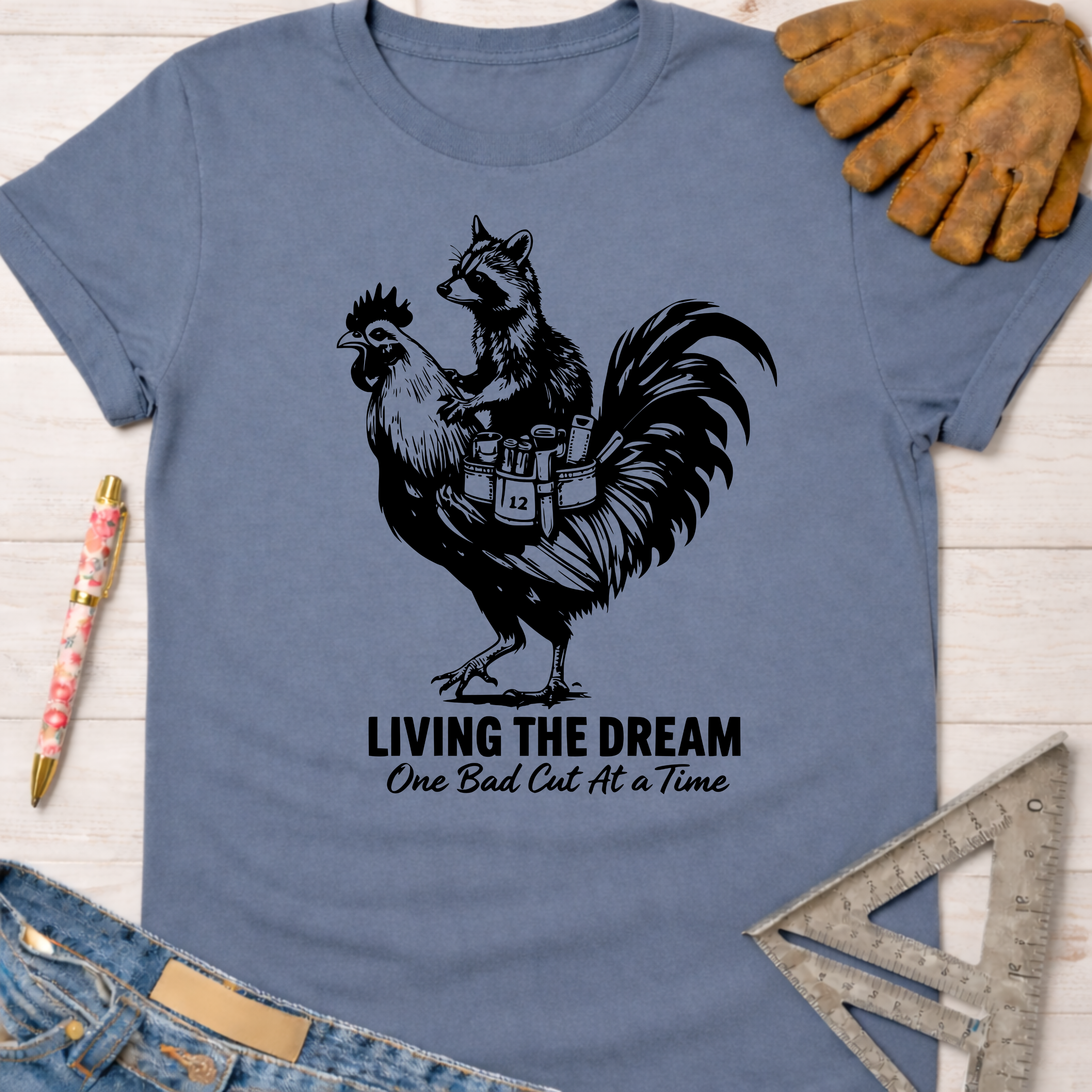Women's funny woodworking graphic tee shirt for female woodworkers, makers, and DIY builders — Sawdust & Ponytails. Blue unisex tee with a raccoon riding a rooster, "Living The Dream One Bad Cut At A Time" text.