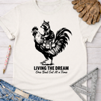 Women's funny woodworking graphic tee shirt for female woodworkers, makers, and DIY builders — Sawdust & Ponytails. White unisex tee with a raccoon riding a rooster, "LIVING THE DREAM One Bad Cut At a Time" text.