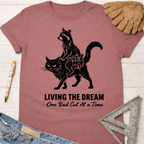 Women's funny woodworking graphic tee shirt for female woodworkers, makers, and DIY builders — Sawdust & Ponytails. Mauve cotton tee with a raccoon riding a cat graphic and "Living The Dream One Bad Cut At A Time" text.