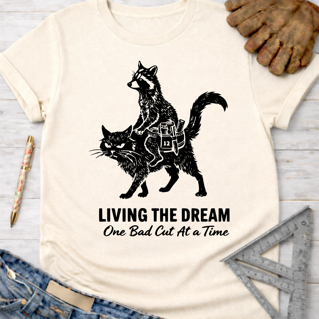 Women's funny woodworking graphic tee shirt for female woodworkers, makers, and DIY builders — Sawdust & Ponytails. Cream tee with a raccoon riding a cat, "LIVING THE DREAM One Bad Cut At A Time" text.