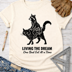 Women's funny woodworking graphic tee shirt for female woodworkers, makers, and DIY builders — Sawdust & Ponytails. Cream tee with a raccoon riding a cat, "LIVING THE DREAM One Bad Cut At A Time" text.