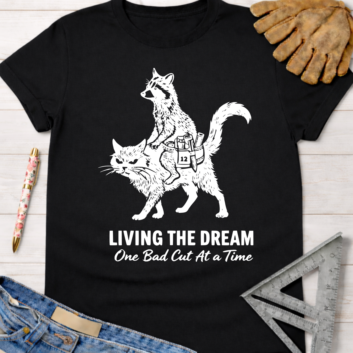 Women's funny black cotton graphic tee with raccoon riding a cat, "Living The Dream One Bad Cut At A Time" text. Sawdust & Ponytails.