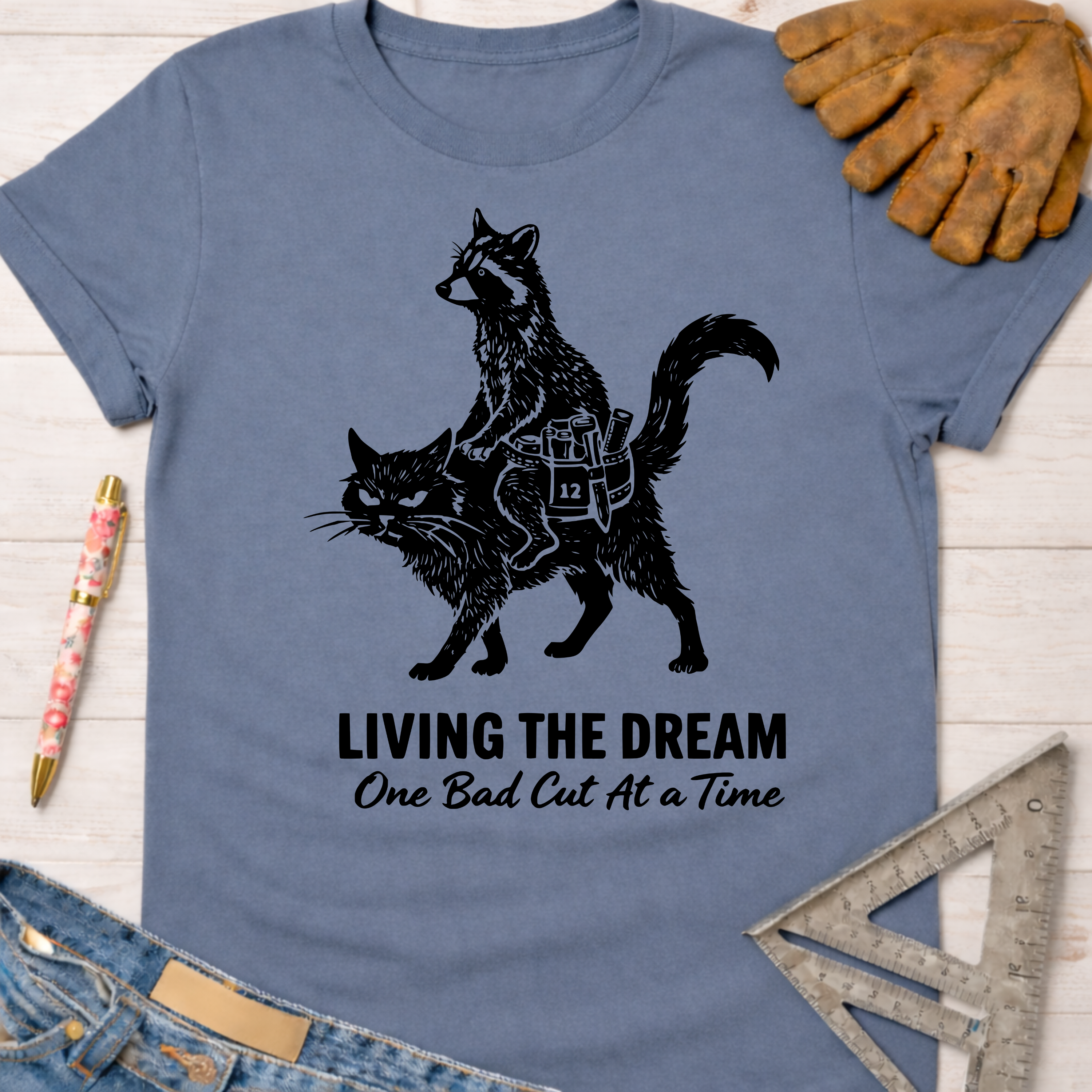 Women's funny woodworking graphic tee shirt for female woodworkers, makers, and DIY builders — Sawdust & Ponytails. Blue cotton tee with a black graphic of a raccoon riding a cat, text "Living The Dream One Bad Cut At A Time.