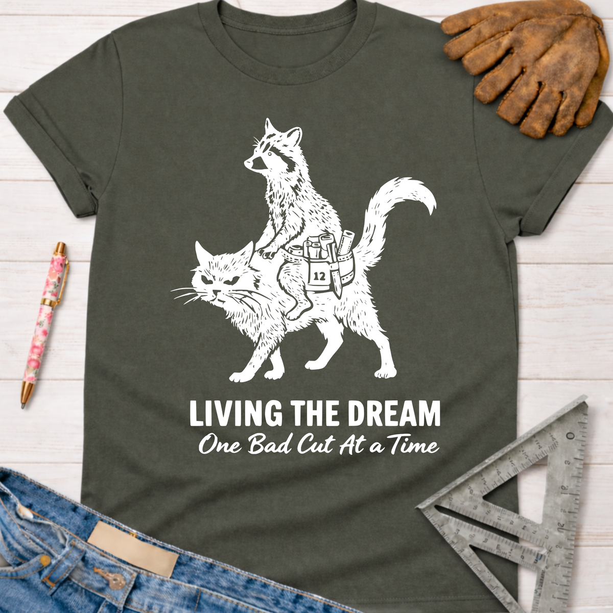 Women's funny woodworking graphic tee shirt for female woodworkers, makers, and DIY builders — Sawdust & Ponytails. Dark gray cotton tee with a white graphic of a raccoon riding a cat, text "LIVING THE DREAM One Bad Cut At A Time".