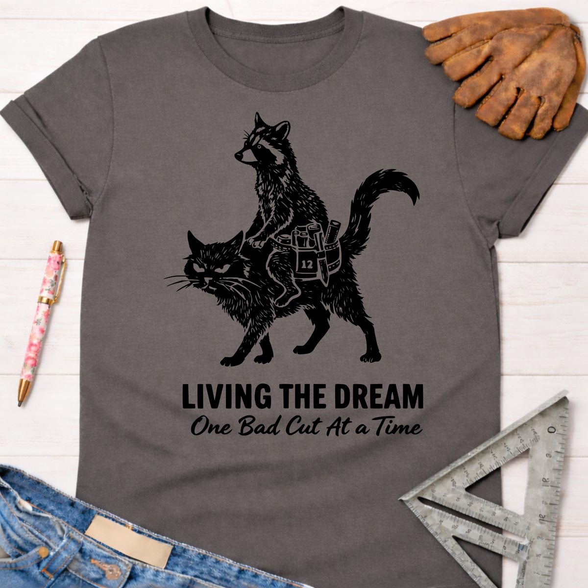 Women's funny charcoal gray tee with a raccoon riding a cat graphic and "Living The Dream One Bad Cut At A Time" text. Sawdust & Ponytails.