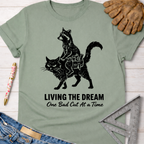 Women's funny woodworking graphic tee shirt for female woodworkers, makers, and DIY builders — Sawdust & Ponytails. Sage green cotton tee with a black graphic of a raccoon riding a cat, text "LIVING THE DREAM One Bad Cut At A Time.