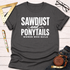 Charcoal gray "Sawdust and Ponytails Women Who Build" T-shirt with white text, flat lay on wood.