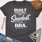 Dark gray women's short-sleeve tee with "Built Something. Got Sawdust. in my BRA." in white text.