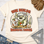 Women's funny woodworking graphic tee shirt for female woodworkers, makers, and DIY builders — Sawdust & Ponytails. Cream t-shirt with a cartoon highland cow building wood, text "SHE BUILDS BEAUTIFUL THINGS".