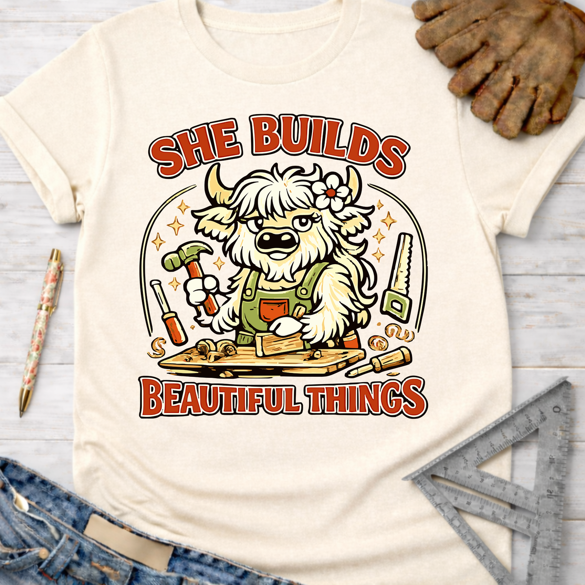 Women's funny woodworking graphic tee shirt for female woodworkers, makers, and DIY builders — Sawdust & Ponytails. Cream t-shirt with a cartoon highland cow building wood, text "SHE BUILDS BEAUTIFUL THINGS".