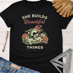 Black cotton tee with "She Builds Beautiful Things" graphic, miter saw, and flowers. Women's funny woodworking graphic tee shirt for female woodworkers, makers, and DIY builders — Sawdust & Ponytails.