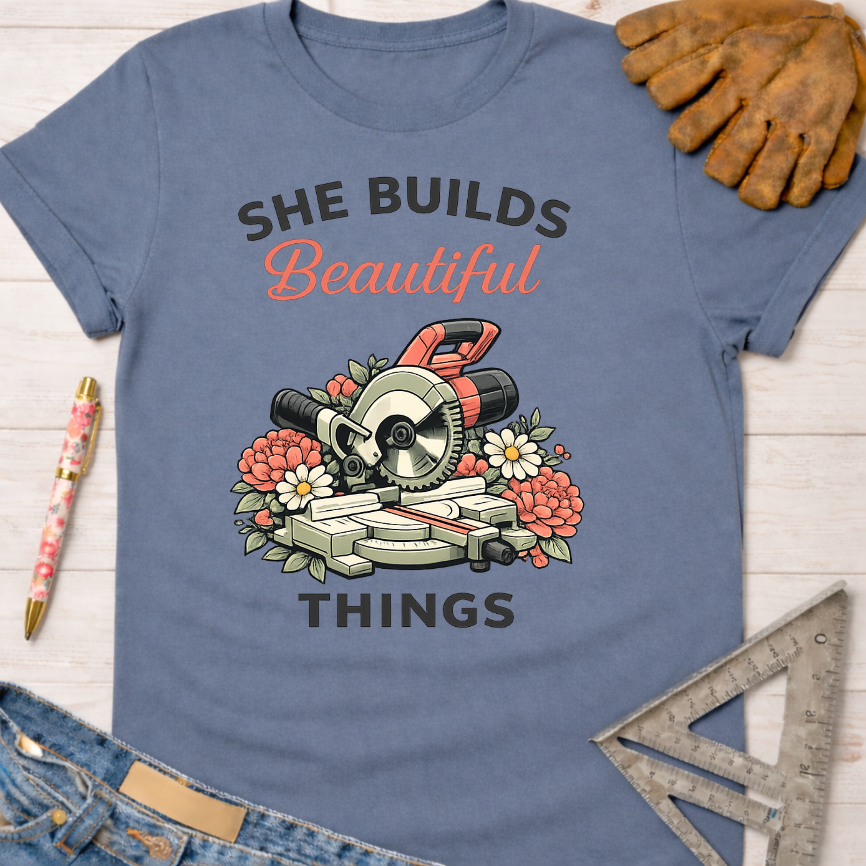 Women's funny woodworking graphic tee shirt for female woodworkers, makers, and DIY builders — Sawdust & Ponytails. Blue cotton t-shirt with "She Builds Beautiful Things" text and a miter saw surrounded by flowers.