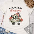 Women's funny woodworking graphic tee shirt for female woodworkers, makers, and DIY builders — Sawdust & Ponytails. White cotton tee with "She Builds Beautiful Things" text and a miter saw surrounded by flowers.