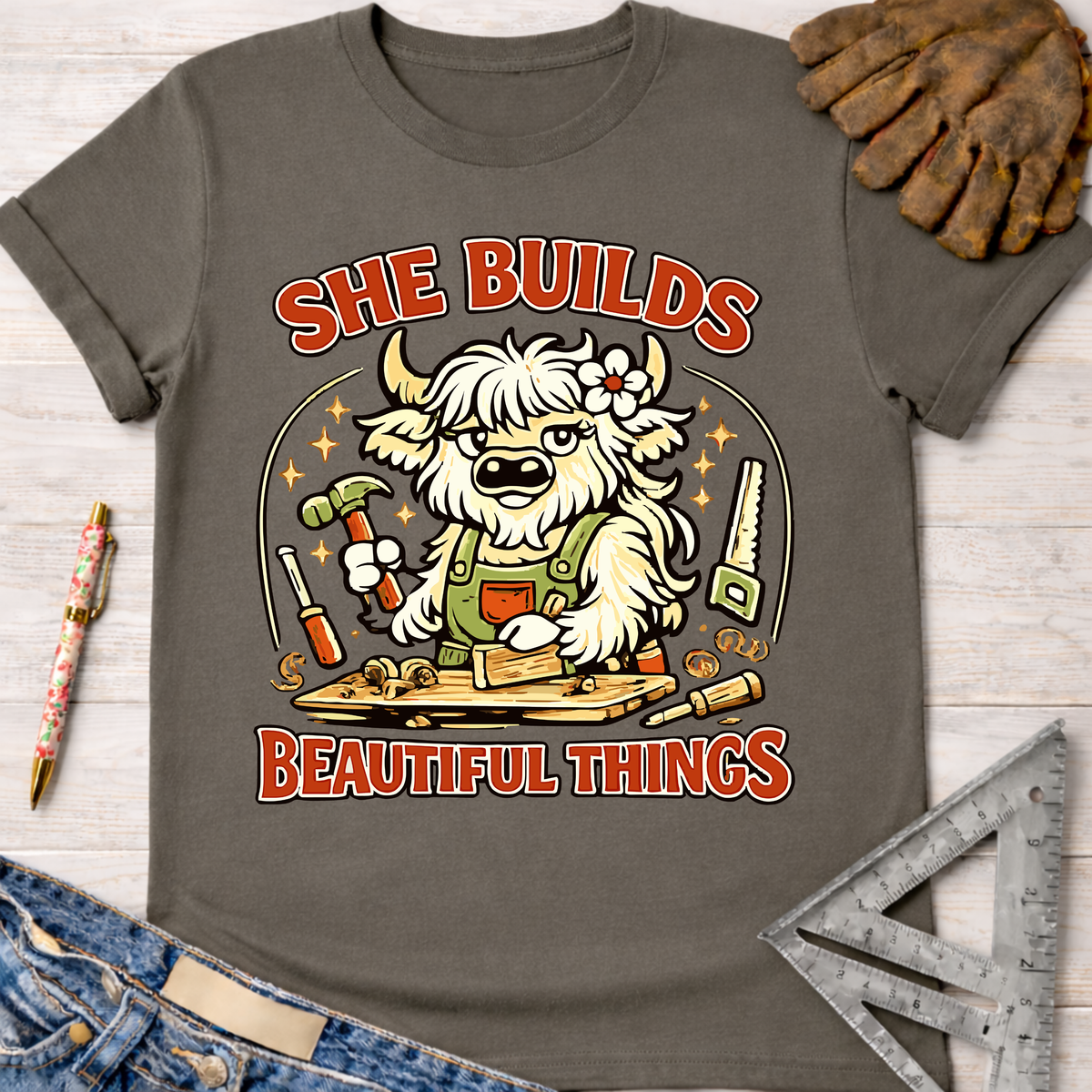 Women's funny woodworking graphic tee shirt for female woodworkers, makers, and DIY builders — Sawdust & Ponytails. Dark gray tee with a cartoon highland cow building wood, text "She Builds Beautiful Things.