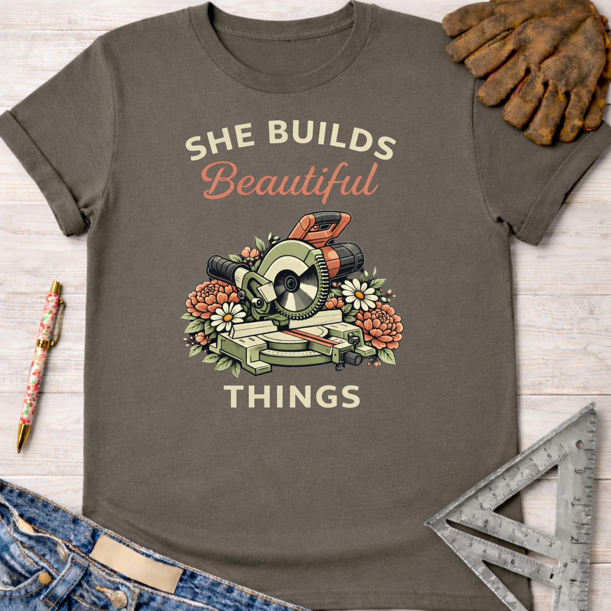 Women's funny woodworking graphic tee shirt for female woodworkers, makers, and DIY builders — Sawdust & Ponytails. Dark gray t-shirt with "She Builds Beautiful Things" text and a miter saw surrounded by flowers.