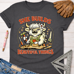Charcoal gray cotton tee with a cartoon highland cow building wood, "She Builds Beautiful Things" text. Women's funny woodworking graphic tee shirt for female woodworkers, makers, and DIY builders — Sawdust & Ponytails