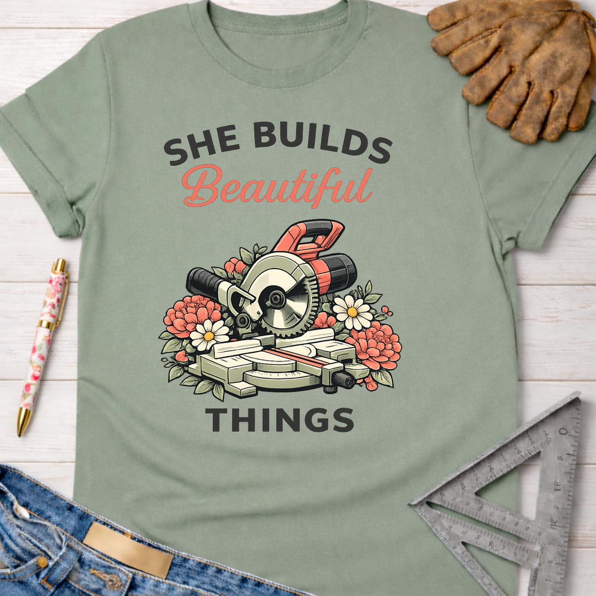 Women's funny woodworking graphic tee shirt for female woodworkers, makers, and DIY builders — Sawdust & Ponytails. Sage green cotton tee with "She Builds Beautiful Things" text and miter saw surrounded by flowers.