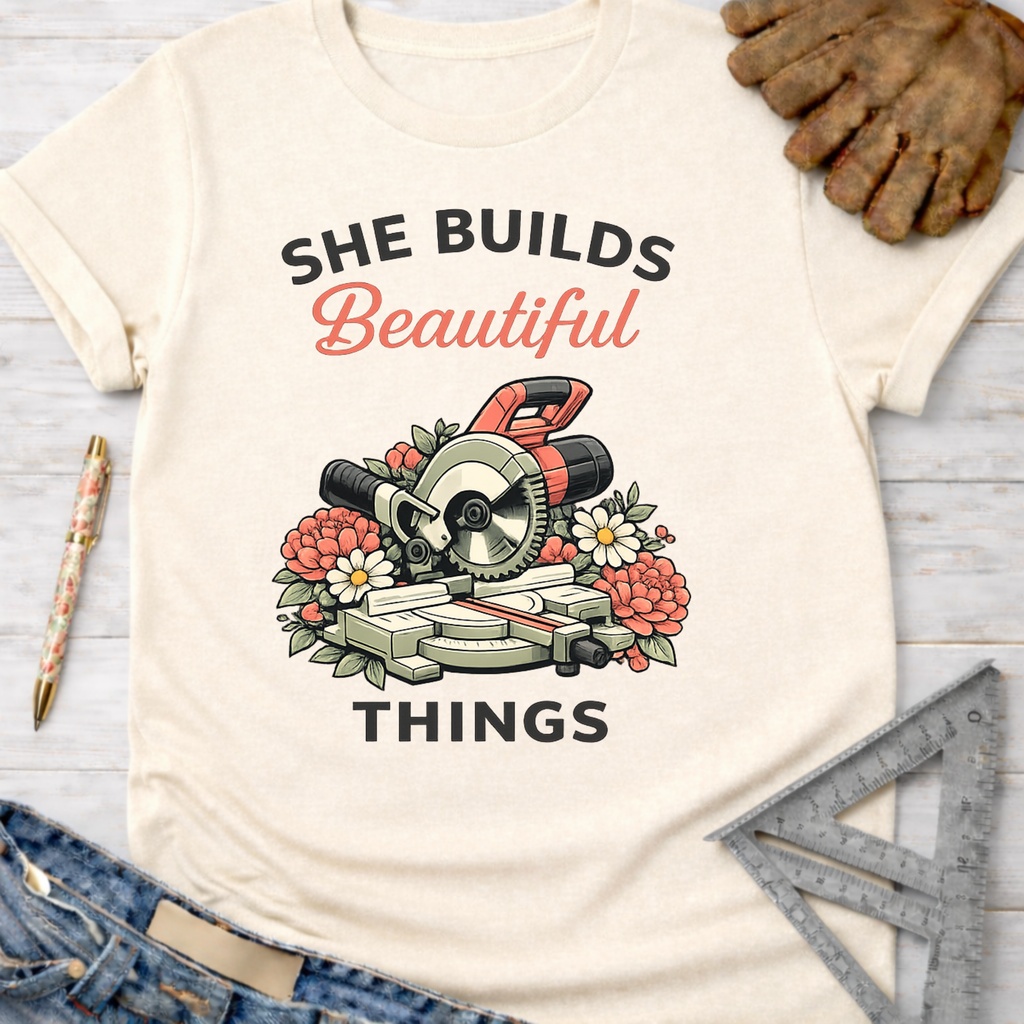 Women's funny woodworking graphic tee shirt for female woodworkers, makers, and DIY builders — Sawdust & Ponytails. Cream t-shirt with "She Builds Beautiful Things" text and a miter saw surrounded by flowers.