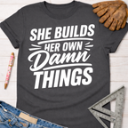 Charcoal gray women's "She Builds Her Own Damn Things" graphic tee, with work gloves and tools.