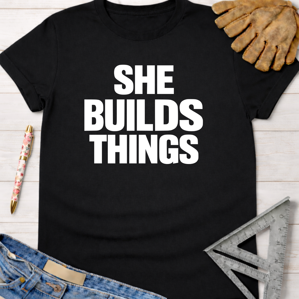 Black cotton "SHE BUILDS THINGS" graphic tee for women, with work gloves, jeans, and a ruler. Sawdust & Ponytails.