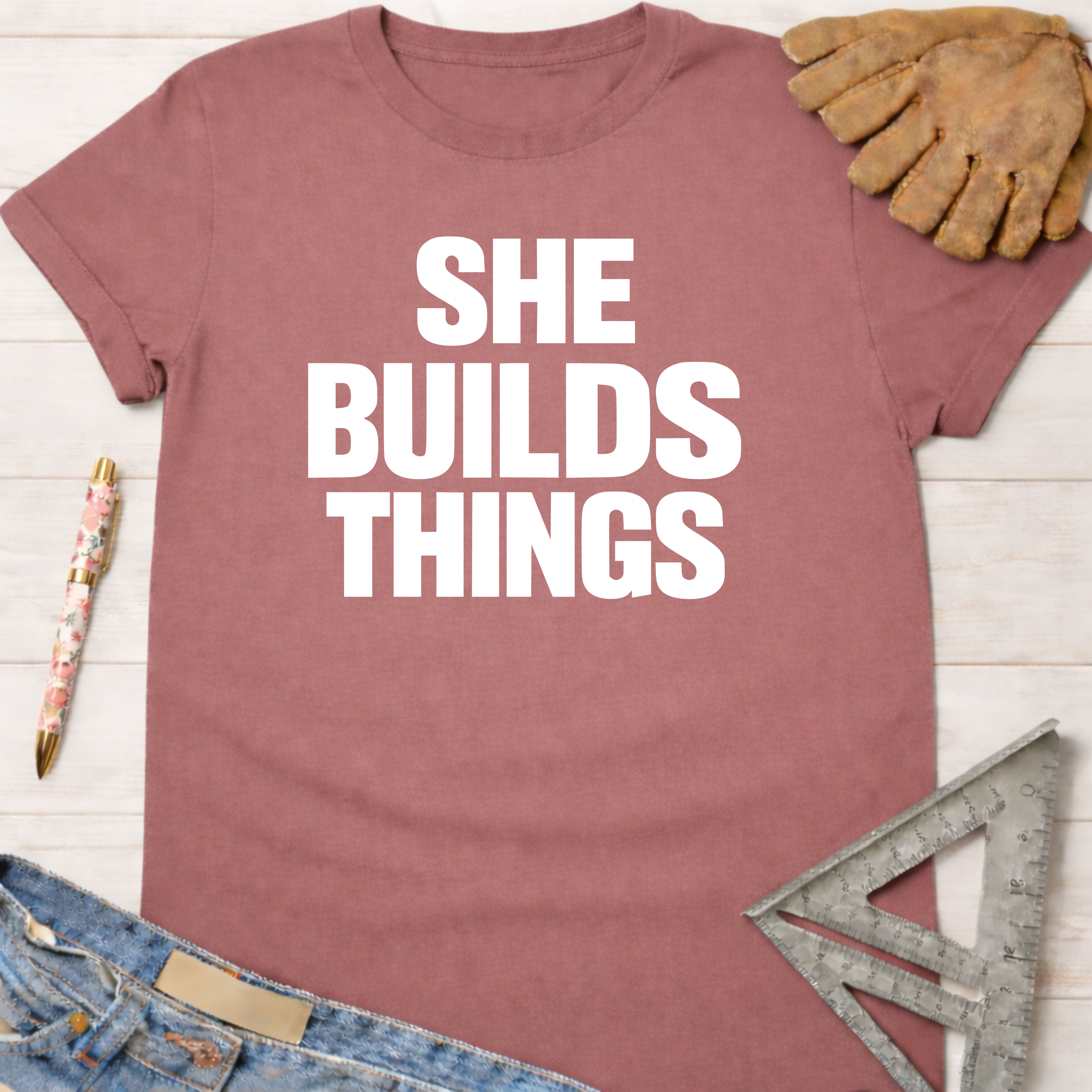 Women's funny woodworking graphic tee shirt for female woodworkers, makers, and DIY builders — Sawdust & Ponytails. Mauve cotton t-shirt with "SHE BUILDS THINGS" in white text, styled with jeans, work glove, and ruler.
