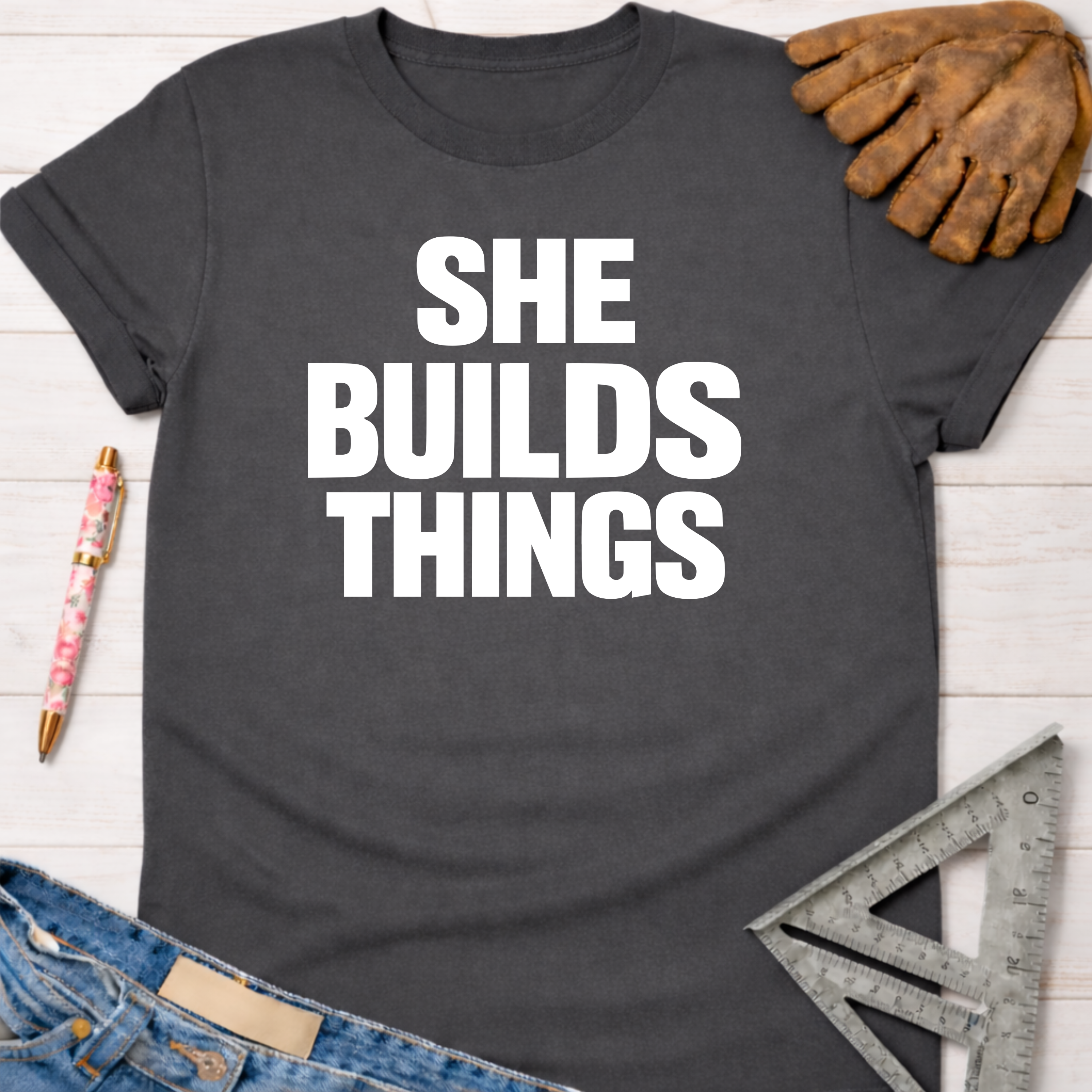 Charcoal grey cotton t-shirt with "SHE BUILDS THINGS" in white text, next to work gloves and a square. Women's funny woodworking graphic tee shirt for female woodworkers, makers, and DIY builders — Sawdust & Ponytails