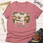 She Builds T-shirt: Dusty rose tee with a graphic of a table saw surrounded by pink flowers and "She Builds." text.