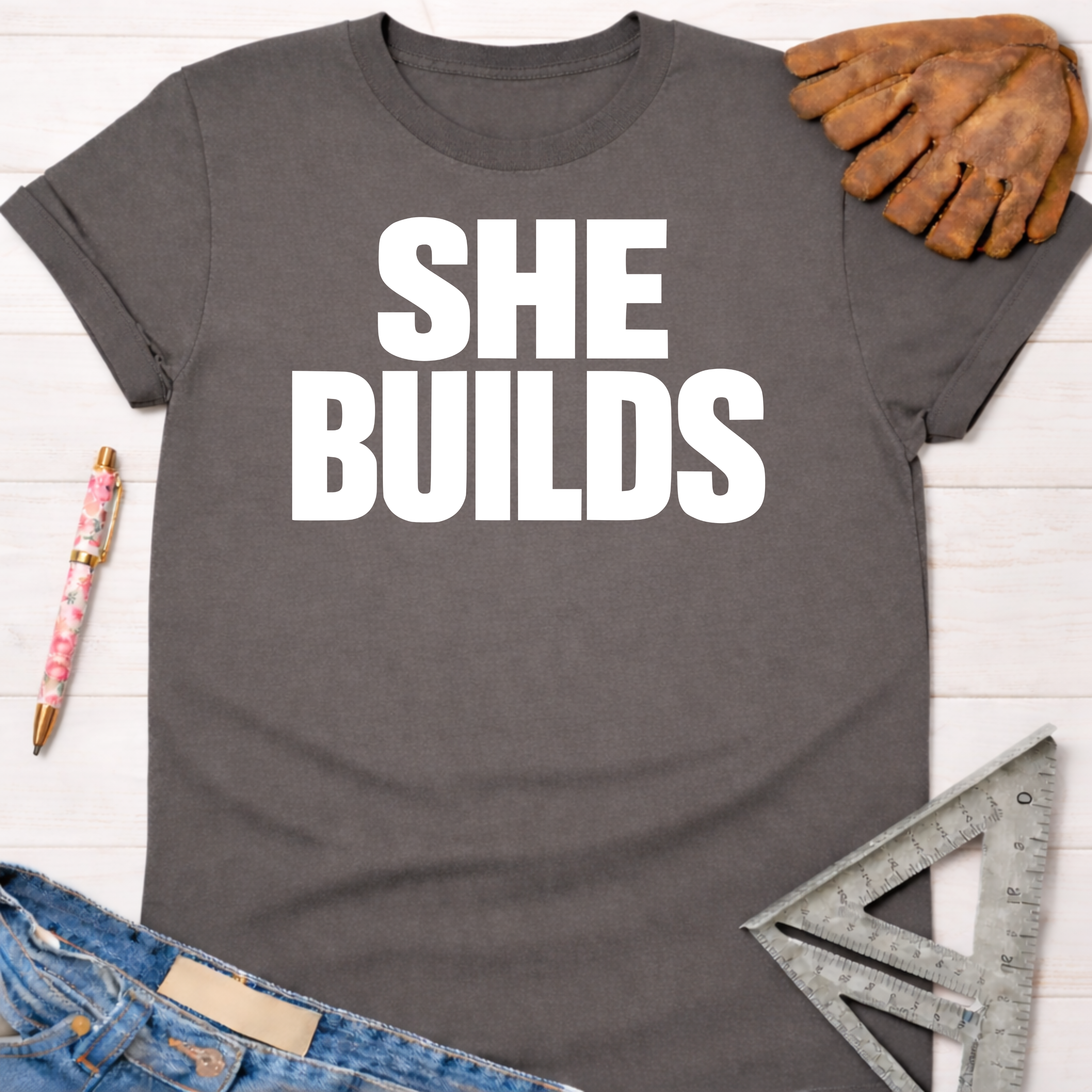 Women's funny woodworking graphic tee shirt for female woodworkers, makers, and DIY builders — Sawdust & Ponytails. Dark gray "SHE BUILDS" t-shirt with rolled sleeves, next to a work glove and jeans.