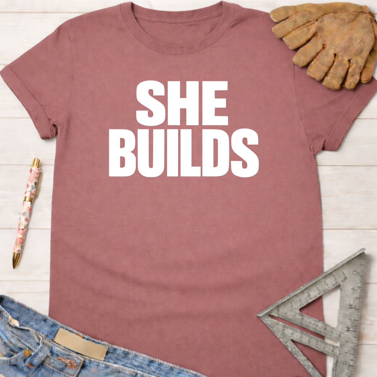 Mauve "SHE BUILDS" graphic tee with work gloves, jeans, pen, and speed square. Women's funny woodworking graphic tee shirt for female woodworkers, makers, and DIY builders — Sawdust & Ponytails