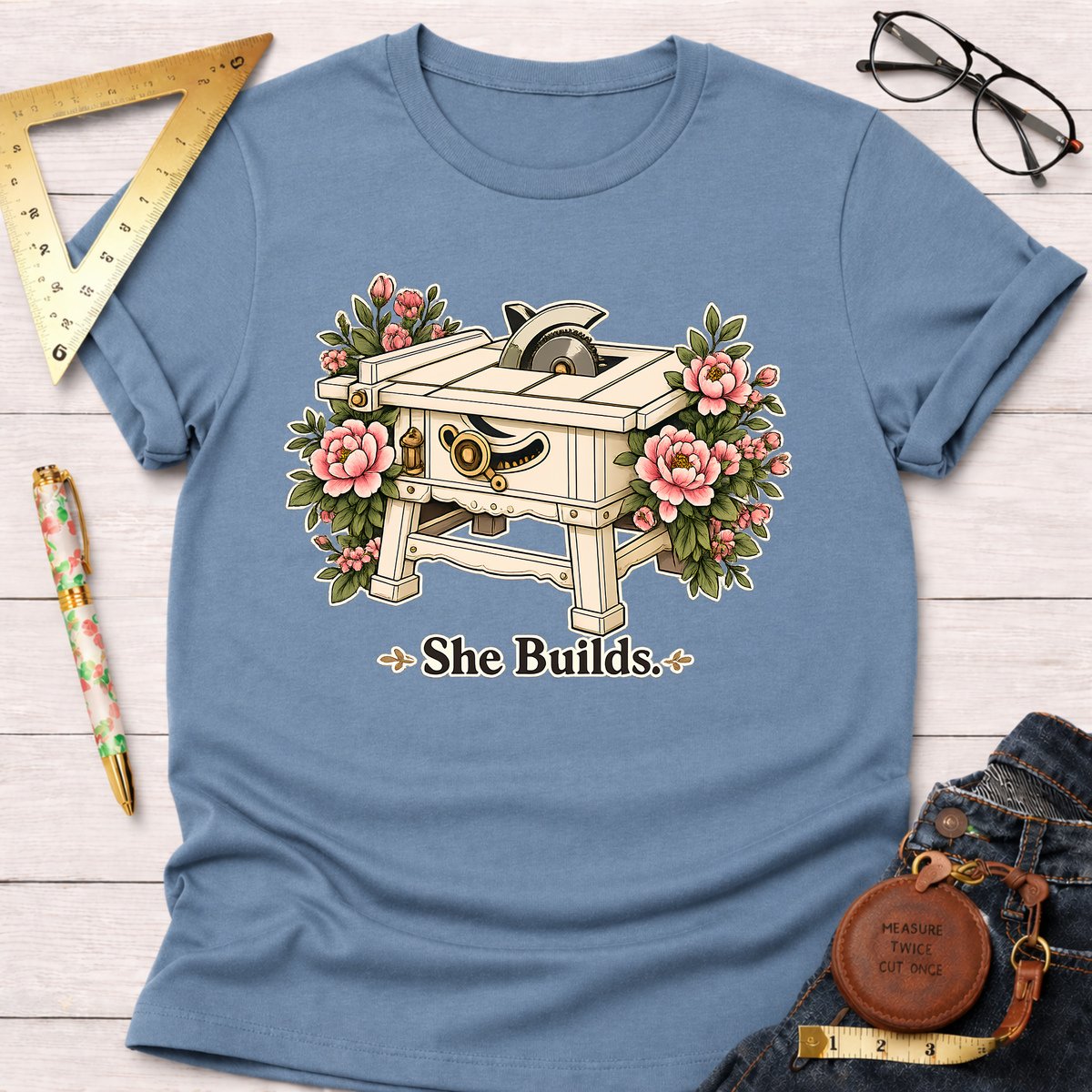 Light blue "She Builds" t-shirt with a table saw and pink floral design.