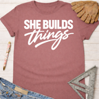 Women's funny woodworking graphic tee shirt for female woodworkers, makers, and DIY builders — Sawdust & Ponytails. Mauve cotton t-shirt with "SHE BUILDS things" in white text.
