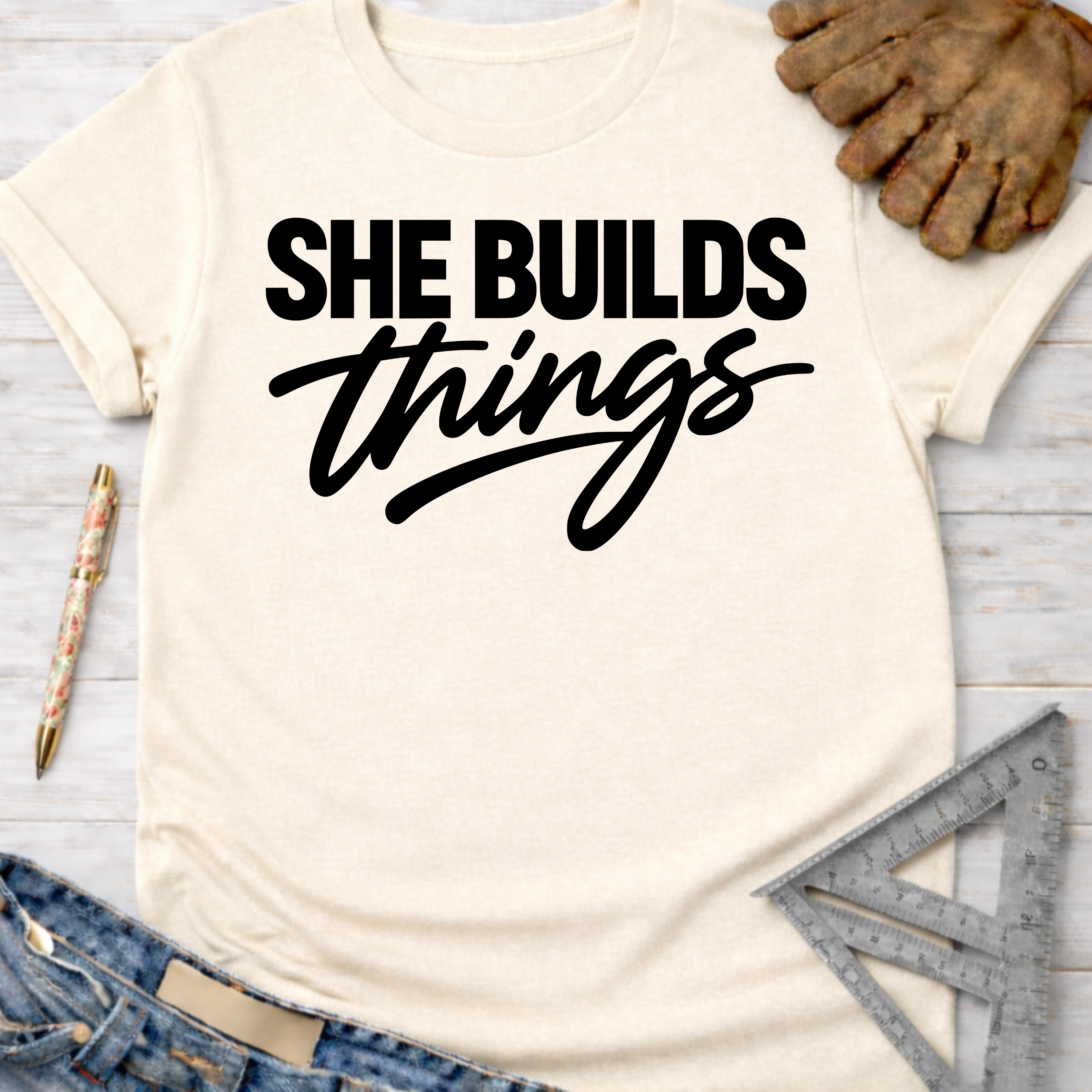 Women's funny woodworking graphic tee shirt for female woodworkers, makers, and DIY builders — Sawdust & Ponytails. Cream t-shirt with "SHE BUILDS things" in black script.