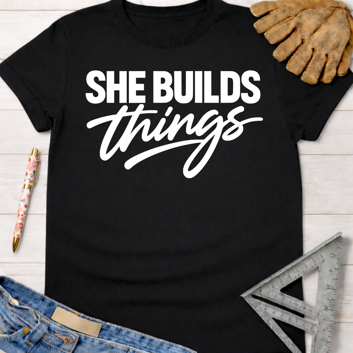 Black cotton "She Builds Things" graphic tee, with work gloves, jeans, and a ruler. Women's funny woodworking graphic tee shirt for female woodworkers, makers, and DIY builders — Sawdust & Ponytails.