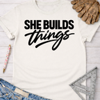 Women's funny woodworking graphic tee shirt for female woodworkers, makers, and DIY builders — Sawdust & Ponytails. Cream short-sleeve cotton tee with "SHE BUILDS things" in black text.