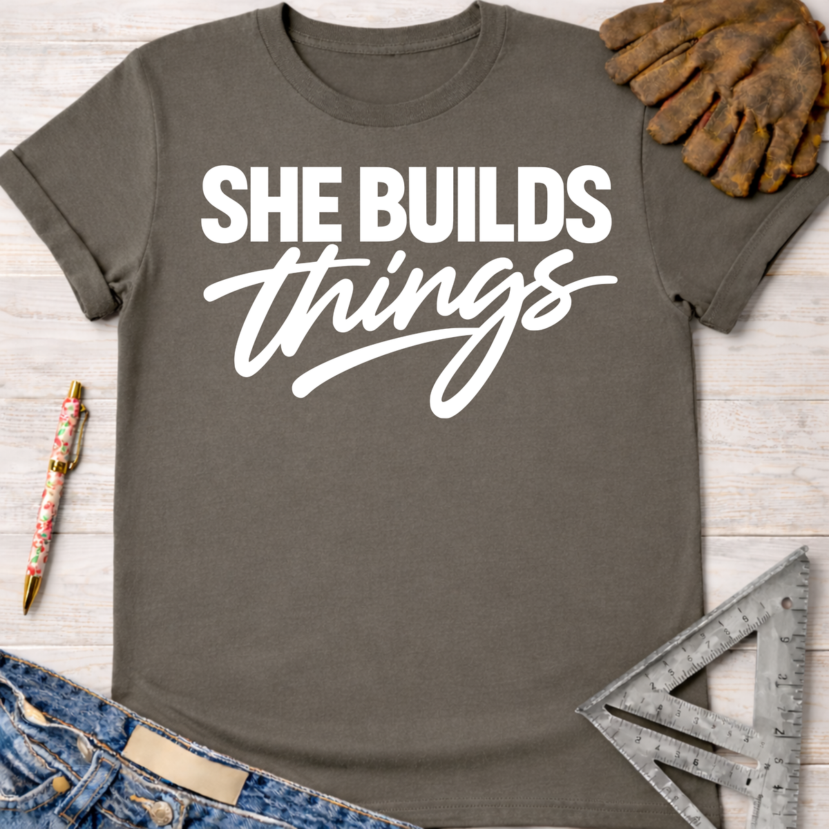 Charcoal grey women's "She Builds Things" graphic tee, with work gloves, jeans, and a ruler.
