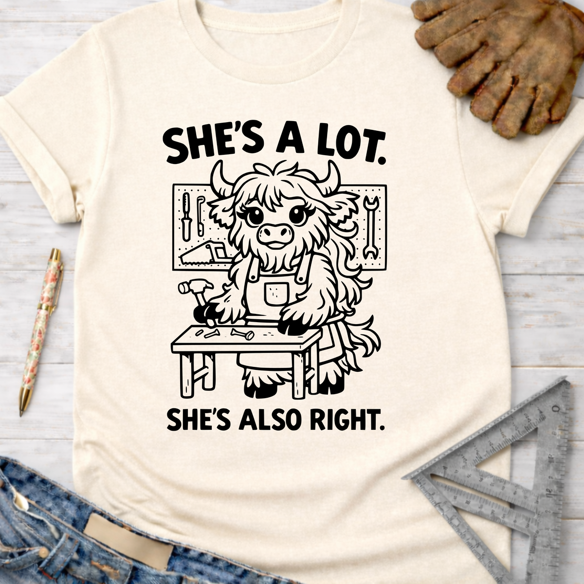 Women's funny woodworking graphic tee shirt for female woodworkers, makers, and DIY builders — Sawdust & Ponytails. Cream t-shirt with "She's A Lot. She's Also Right." text and a cute highland cow working at a workbench.