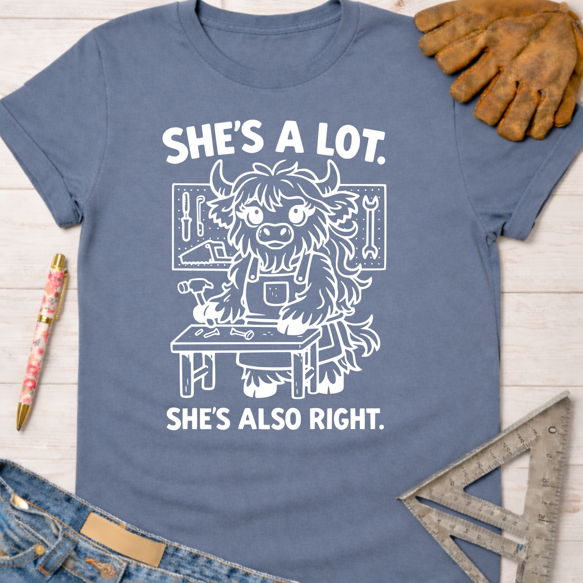 Women's funny woodworking graphic tee shirt for female woodworkers, makers, and DIY builders — Sawdust & Ponytails. Blue cotton tee with "She's A Lot. She's Also Right." text and a cartoon highland cow builder.