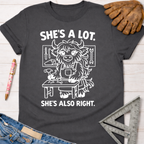 Women's funny woodworking graphic tee shirt for female woodworkers, makers, and DIY builders — Sawdust & Ponytails. Dark gray cotton tee with "She's A Lot. She's Also Right." text and a cartoon highland cow at a workbench.