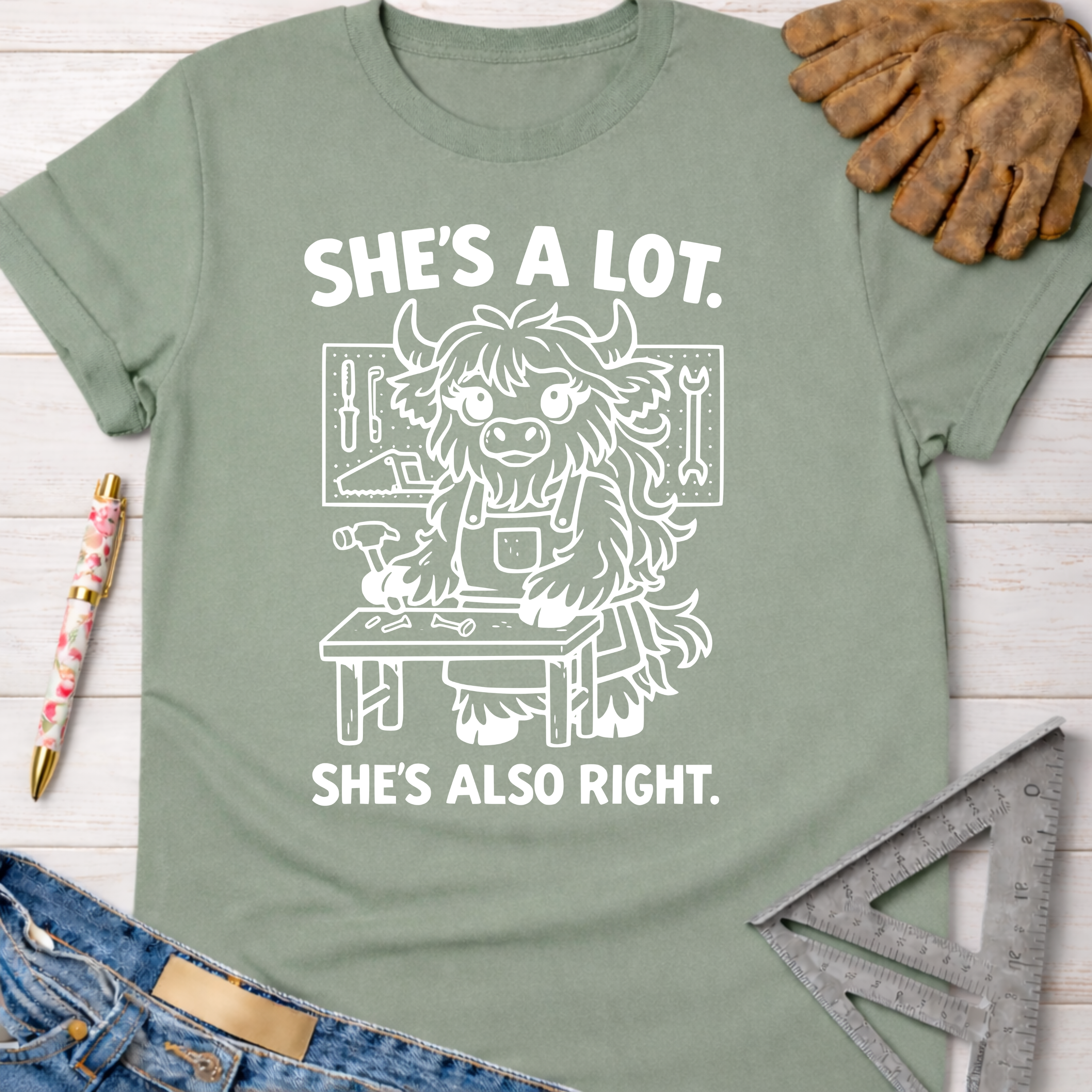 Women's funny woodworking graphic tee shirt for female woodworkers, makers, and DIY builders — Sawdust & Ponytails. Sage green cotton tee with "She's A Lot. She's Also Right." text and a cartoon highland cow building.