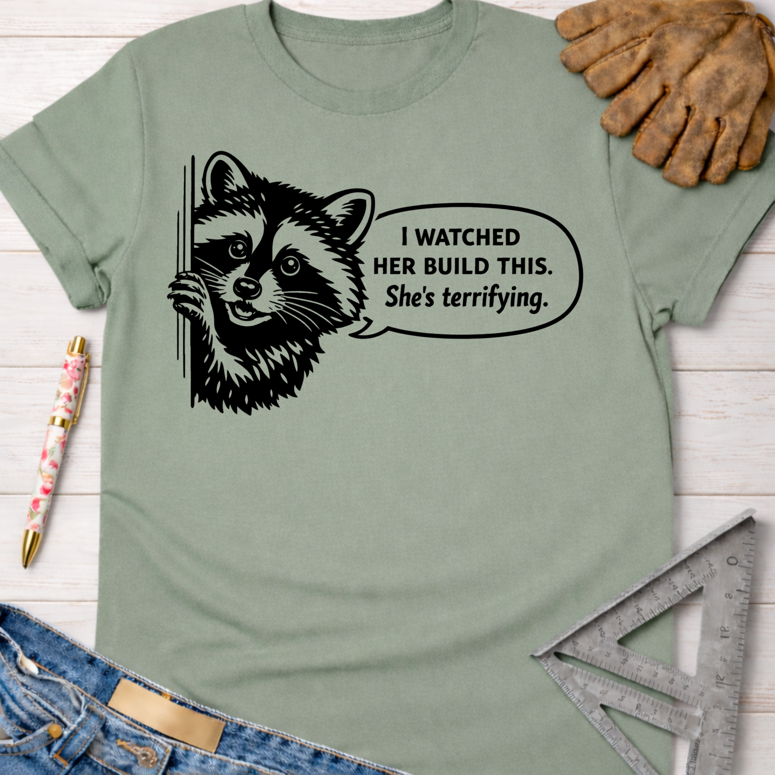 Women's funny woodworking graphic tee shirt for female woodworkers, makers, and DIY builders — Sawdust & Ponytails. Sage green cotton t-shirt with a black graphic of a raccoon and speech bubble: "I watched her build this. She's terrifying.