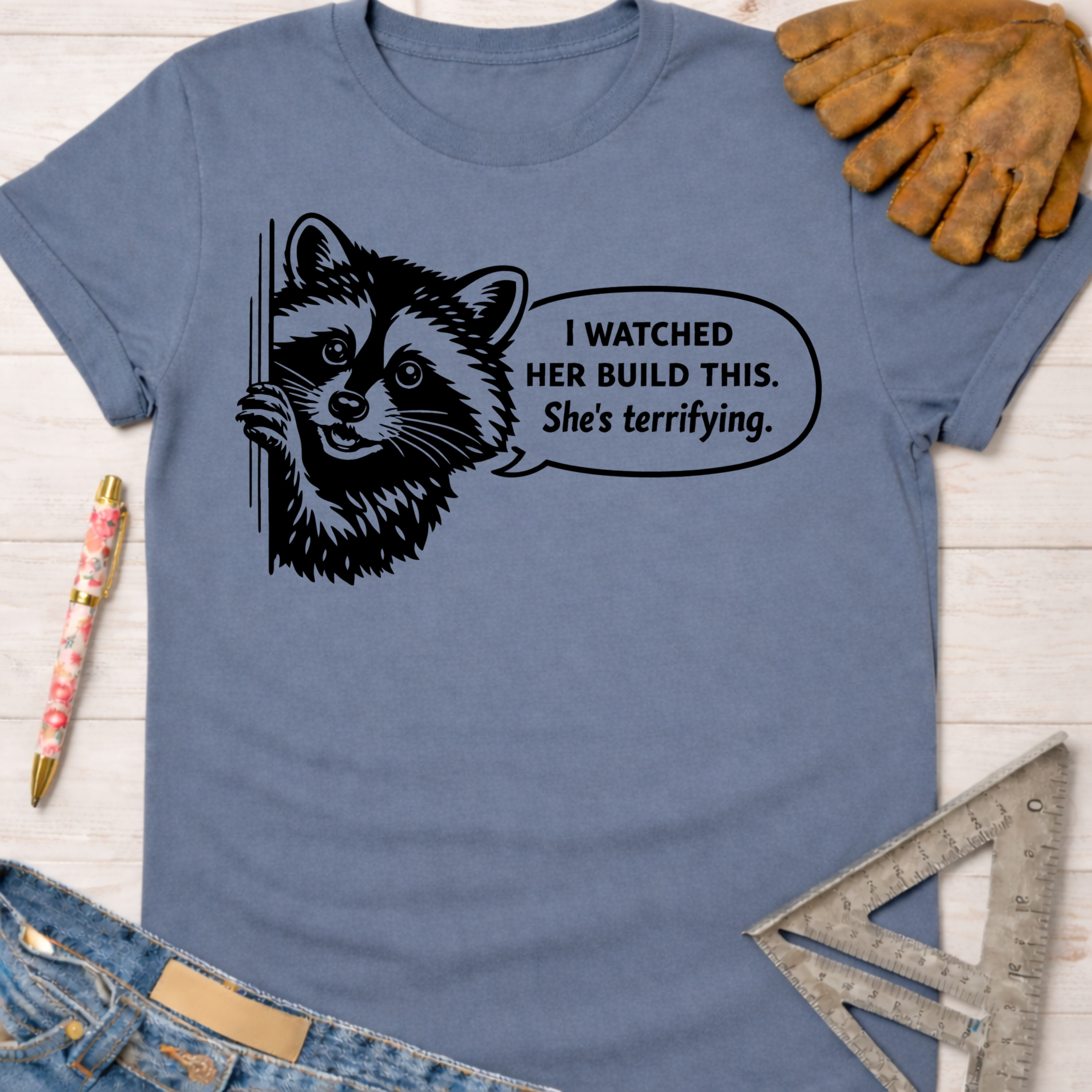 Women's funny woodworking graphic tee shirt for female woodworkers, makers, and DIY builders — Sawdust & Ponytails. Blue cotton t-shirt with a black raccoon graphic and "I watched her build this. She's terrifying." text.