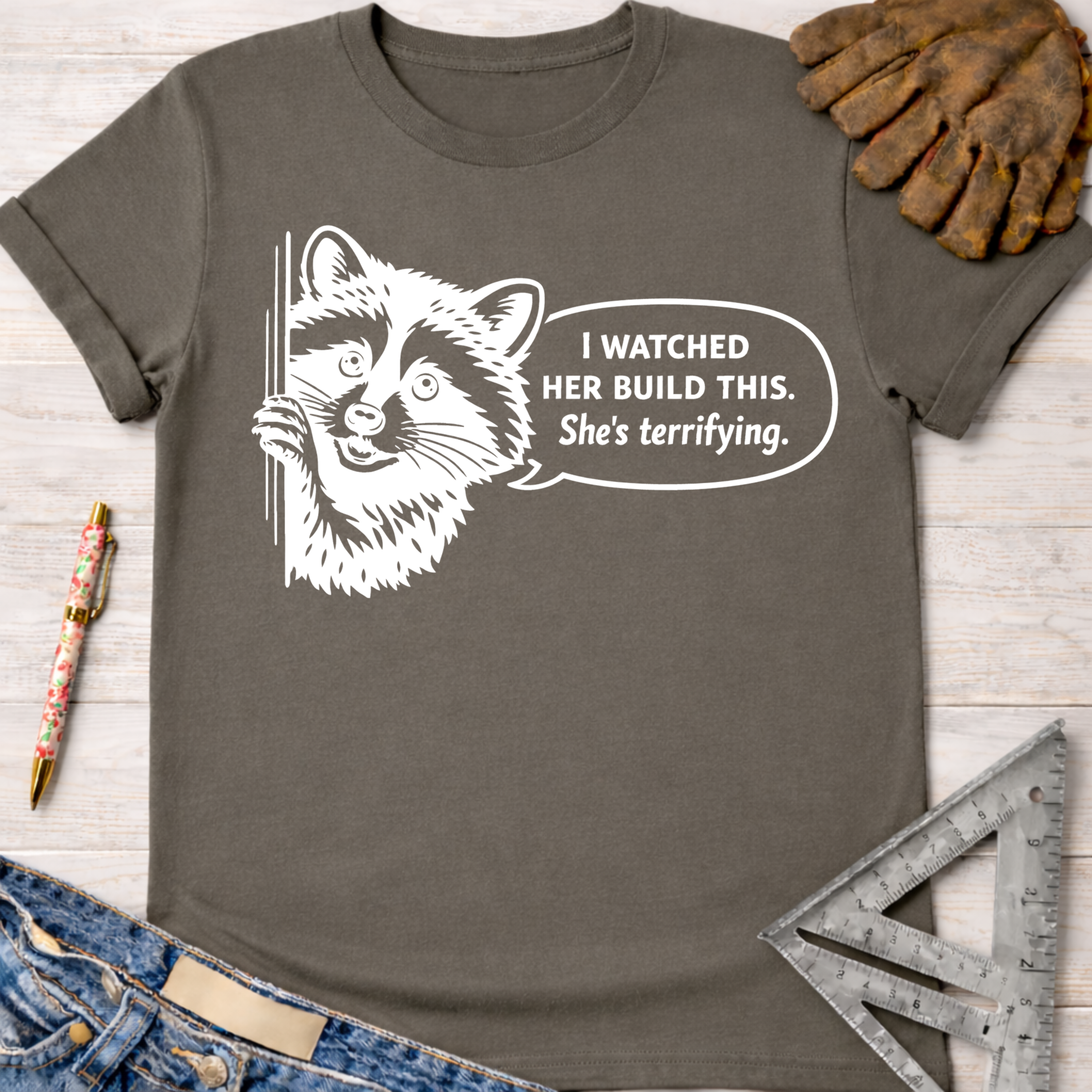 Women's funny woodworking graphic tee shirt for female woodworkers, makers, and DIY builders — Sawdust & Ponytails. Dark gray t-shirt with a white graphic of a raccoon saying "I watched her build this. She's terrifying.