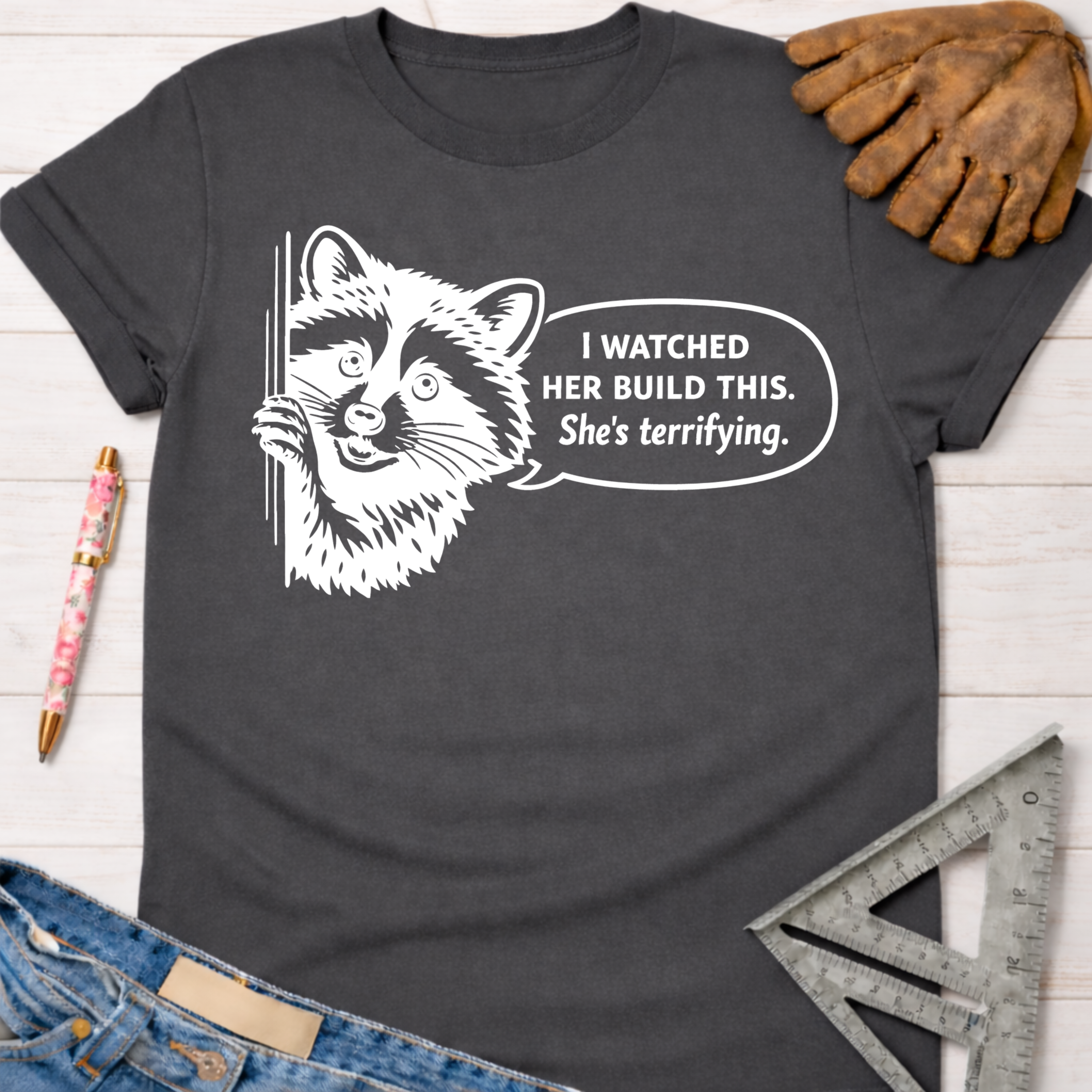 Women's funny woodworking graphic tee shirt for female woodworkers, makers, and DIY builders — Sawdust & Ponytails. Dark gray t-shirt with a white graphic of a raccoon and speech bubble "I watched her build this. She's terrifying.
