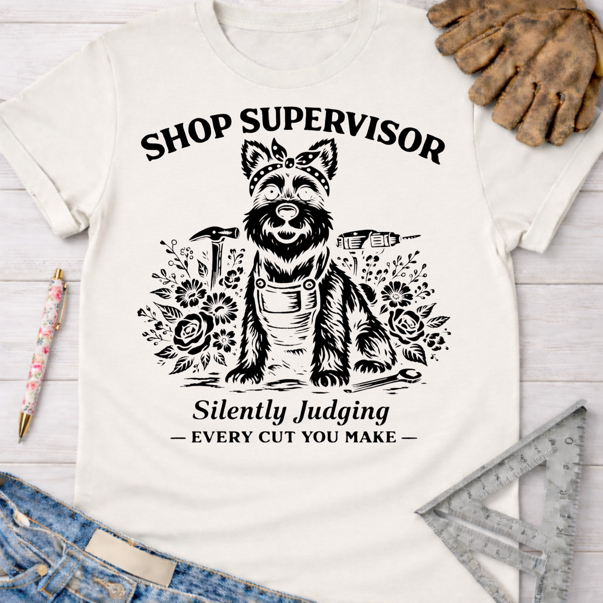 Women's funny woodworking graphic tee shirt for female woodworkers, makers, and DIY builders — Sawdust & Ponytails. White cotton tee with a black graphic of a dog in a bandana and apron, surrounded by tools and flowers, with text "Shop Supervisor Silently Judging Every Cut You Make.